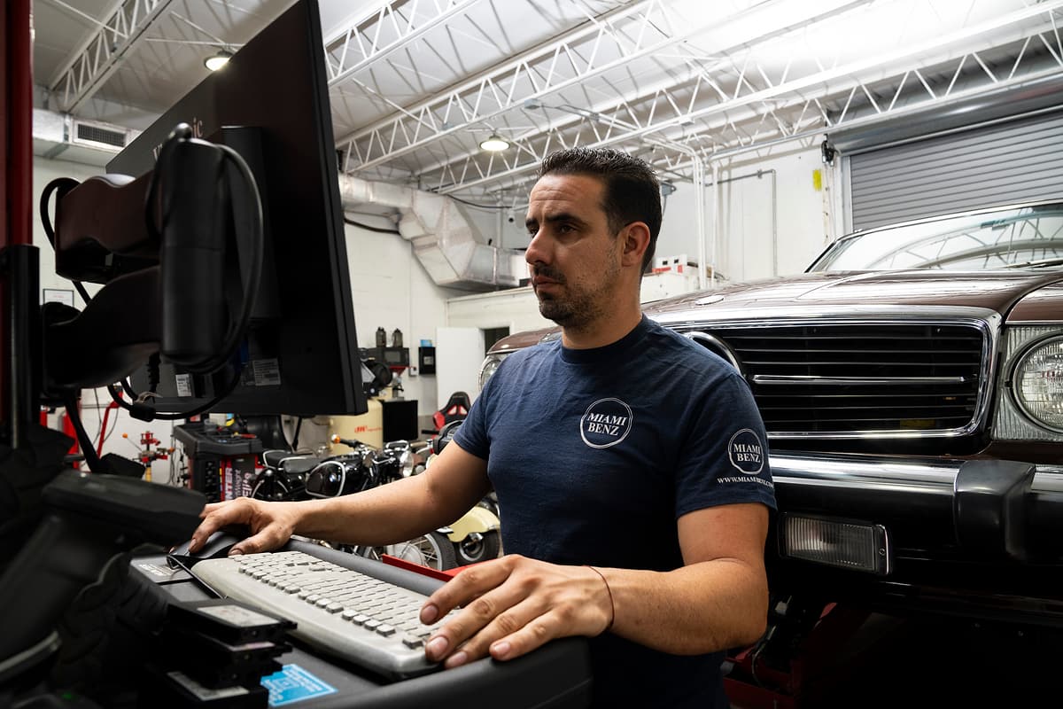 Miami Benz technician working at the workshop diagnostic computer beside a car.