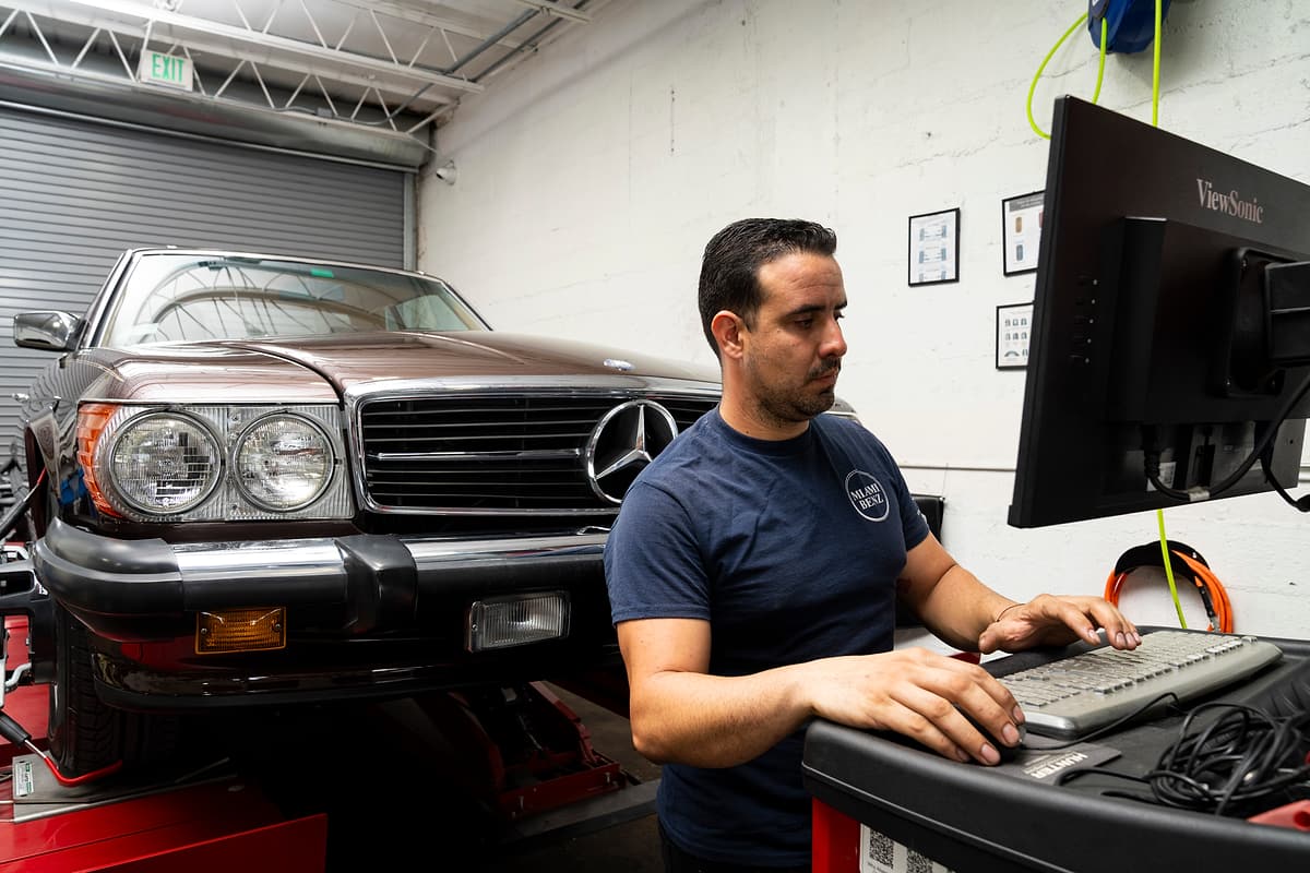 Miami Benz technician at the diagnostic screen with a Mercedes-Benz in the workshop bay behind.